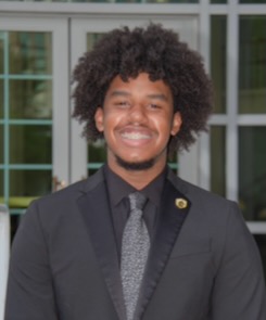 Photo of Deylynd Grant smiling and wearing a dark suit with a black shirt and gray tie standing in front of French doors.