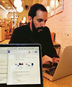 Photo of Nick Smerker staring at a laptop computer, with a separate laptop computer screen facing the camera. Nick is sitting a room painted white and wearing a black sweater.