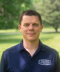 Photo of Corey Gracie-Griffin wearing a dark blue polo shirt with a logo saying "Penn State Fullers Overlook" and standing in front of a leafy green background