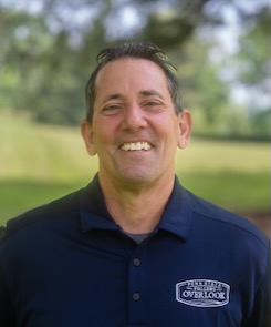 Photo of Joseph Reno wearing a dark blue polo shirt with the "Penn State Fullers Overlook" logo and standing against a shaded, leafy green backdrop