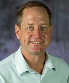 Photo of Travis Lesser wearing a light spearmint green dress shirt and standing against a grey background