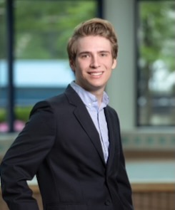 Photo of Julius Camper wearing a dark blue blazer and light blue collared shirt standing in front of some windows with trees in the background