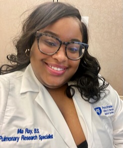 Photo of Mia Ray wearing black glasses and a white lab coat with her name on it and the title of "Pulmonary Research Specialist" as well as a Penn State Hershey logo