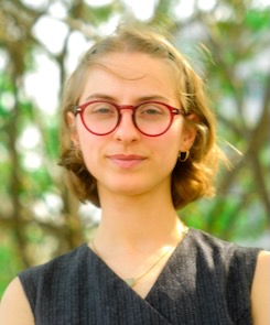 Photo of Emily Weaver wearing a blue and gray striped sleeveless top, pendant necklace, and red glasses while standing in front of trees