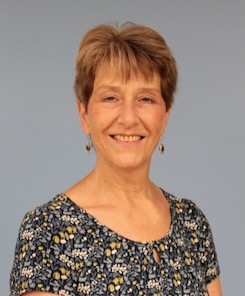 Photo of Jeanette Fornwalt standing in front of a gray background wearing a black top patterned with yellow and white flecks and wearing black drop earrings