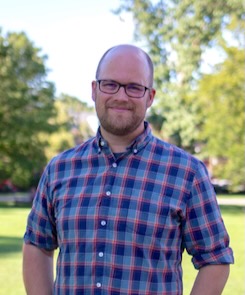Photo of Eric Sauder wearing a blue and purple plaid shirt while standing in front of some trees in a field