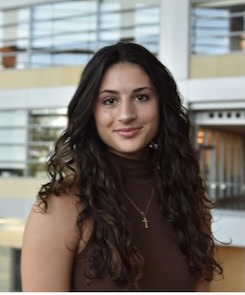 Jessica Padula Headshot of a person with brown bangs and a blue jacket looking at the camera, with a green background.