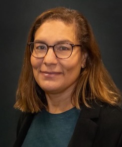 Photo of Coren Jagnow, a woman wearing glasses with light brown shoulder-length hair and wearing a black suit jacket and dark green top